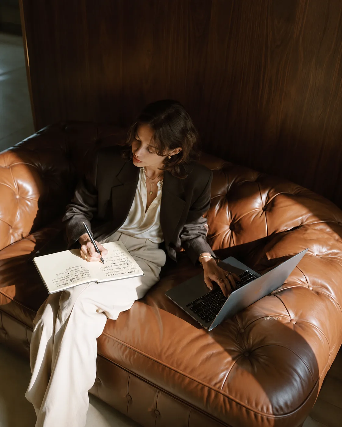 A woman writing in a journal with a laptop on a leather sofa